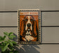 A framed poster of a Basset Hound dog is displayed on a wall, with a plant to the left of the frame.