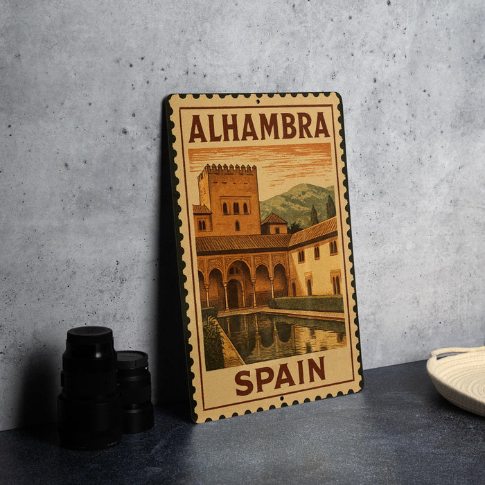 A metal sign with a vintage-style illustration of a castle and a pool, labeled "ALHAMBRA" and "SPAIN", is displayed on a table.