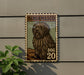 A postage stamp-like sign featuring a large, shaggy dog with the text "BERGAMASCO" and "DOG 20" printed on it, mounted on a wall next to a plant.