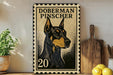 A metal sign featuring a black and tan illustration of a Doberman Pinscher dog, with the text "DOBERMAN PINSCHER" printed on it. The sign is placed on a wooden surface, with a potted plant and a wooden cutting board visible in the background.