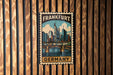 A vintage-style metal stamp depicting the city of Frankfurt, Germany, with a bridge and skyline in the background.