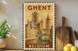 A vintage-style postage stamp from Belgium featuring a cityscape with a river and a bridge, placed on a wooden surface next to a wooden cutting board and a potted plant.
