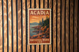 A vintage-style postage stamp depicting a scenic landscape with mountains, pine trees, and rocky cliffs, hanging on a wooden wall.