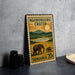 A metal sign with a vintage design, featuring an illustration of elephants and a landscape with mountains, placed on a table next to some black camera lenses.