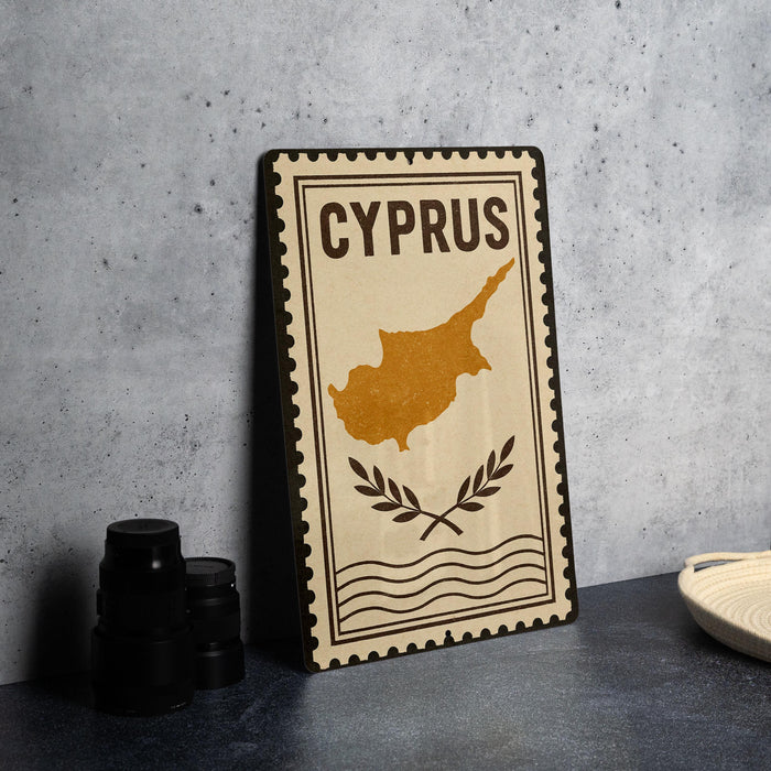 a decorative metal sign with the word "CYPRUS" on it, along with a map of the island of Cyprus. The sign is placed on a table next to some black camera lenses.