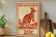 A red kangaroo stamp with the text "RED KANGAROO" and "20c" printed on it, placed on a wooden surface next to a potted plant and a wooden cutting board.