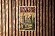 A vintage-style metal sign with the text "PÉCS HUNGARY" displayed prominently in the center, featuring an illustration of a cathedral or church.