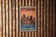 A vintage-style poster of the Pittsburgh skyline, featuring a bridge and the city's name, is displayed on a wooden wall.