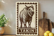a wooden sign with a picture of a buffalo, which is labeled "Takin". The sign is placed on a shelf next to a potted plant and a wooden cutting board.