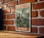 A vintage-style poster of La Gomera mountain in Spain, featuring a lush green landscape with palm trees and a winding path, is displayed on a brick wall.