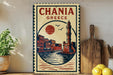 A vintage-style metal sign with the text "CHANIA GREECE" and an illustration of a lighthouse and buildings, placed on a wooden surface next to a potted plant and a cutting board.