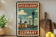 A vintage-style metal sign with a colorful illustration of Düsseldorf, Germany, featuring a tower and a bridge, is displayed on a shelf next to a wooden cutting board and a potted plant.