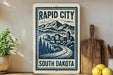 a sign for Rapid City, South Dakota, with a mountain landscape and a road leading to a city.