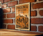 A vintage-style poster of Phoenix, Arizona, USA, featuring a desert landscape with mountains and buildings, mounted on a brick wall.