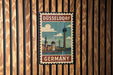 A vintage-style metal stamp with the text "DÜSSELLORF GERMANY" and an illustration of a city skyline, including a tall tower, is displayed on a wooden wall.