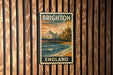 A vintage-style metal sign with the text "BRIGHTON ENGLAND" displayed prominently, featuring a scenic image of a coastal city with a pier and buildings.