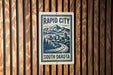 A sign for Rapid City, South Dakota, featuring a mountain landscape and a winding road.