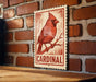 A red cardinal perched on a branch is displayed on a wooden surface against a brick wall.