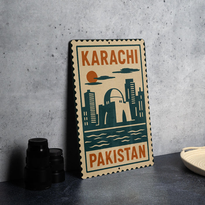 A metal sign with the text "KARACHI PAKISTAN" and an illustration of a city skyline, placed on a gray countertop next to some black camera lenses.