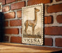 a wooden table with a framed illustration of a llama on it, mounted on a brick wall.