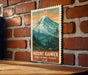 A vintage-style metal sign depicting a mountain landscape with pine trees and a snow-capped peak, mounted on a brick wall.