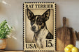 A metal sign featuring a black and white illustration of a Rat Terrier dog, with the text "RAT TERRIER" and "USA 15¢" printed on it. The sign is placed on a wooden surface, with a potted plant, a wooden cutting board, and some lemons nearby.