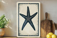 a framed artwork of a starfish on a shelf, with a wooden cutting board and a potted plant nearby.