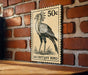 a 50c postage stamp with a drawing of a bird, specifically a Secretary Bird, mounted on a brick wall.