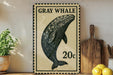 a framed illustration of a gray whale, a 20-cent stamp, and some kitchen utensils on a wooden surface.