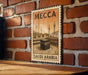 A vintage-style metal sign with the text "MCCA SAUDI ARABIA" and an illustration of the Kaaba, the most sacred site in Islam, is displayed on a brick wall.
