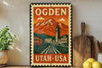 A vintage-style metal sign with the text "OGDEN UTAH USA" and an illustration of a mountain and city, placed on a wooden surface next to a wooden cutting board and a potted plant.