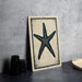 a framed picture of a starfish on a wall, with a stack of black camera lenses and a woven basket placed next to it.