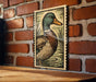 A framed illustration of a duck is hanging on a brick wall.