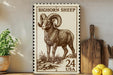 a large stamp-like artwork depicting a bighorn sheep, with the text "BIGHORN SHEEP" and "24 USA" printed on it. The artwork is displayed on a wooden surface, with a potted plant, a wooden cutting board, and some lemons nearby.