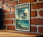 A vintage-style poster of a tropical beach scene with palm trees and the ocean, mounted on a brick wall.