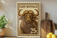a large stamp-like artwork depicting a buffalo, with the text "CAPE BUFFALO" and the denomination "25c" printed on it. The artwork is displayed on a shelf, surrounded by various kitchen utensils and a potted plant.