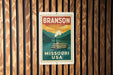 A vintage-style metal sign with the text "BRANSON MISSOURI USA" displayed on a wooden background.
