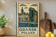 A vintage-style metal sign with a drawing of a cityscape and the text "Gdansk Poland" is displayed on a wooden shelf, accompanied by a potted plant, a wooden cutting board, and some lemons.