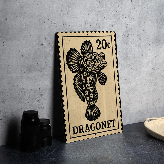 A 20c postage stamp featuring a detailed illustration of a dragonnet fish, placed on a dark surface next to some black camera lenses.