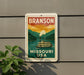 A vintage-style metal sign with a riverboat illustration and the text "BRANSON MISSOURI USA" is displayed on a gray wall.