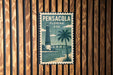 A vintage-style metal sign with the text "PENSACOLA FLORIDA USA" and a lighthouse illustration, mounted on a wooden wall.