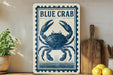 a blue crab on a white sign with a decorative border, placed on a wooden surface next to a wooden cutting board and some lemons.