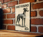 A greyhound dog illustration on a 10c postage stamp is displayed on a brick wall.
