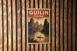 A vintage postage stamp depicting a scenic landscape with mountains, a river, and a person in a boat, hanging on a wooden wall.