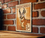 A wooden surface with a wooden frame, and a vintage-style postage stamp featuring a deer on it, mounted on a brick wall.