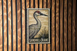 A metal stamp-like sign featuring a sandhill crane is displayed on a wooden wall.