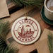 A circular ornament with the text "AARHUS DENMARK" printed on it, surrounded by pine branches and a cup of coffee on a wooden surface.