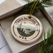 A circular ornament with the text "ACADIA NATIONAL PARK USA" printed on it, surrounded by pine branches and pine needles.