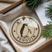 A decorative ornament featuring a penguin is displayed in a box, surrounded by pine branches.