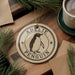 A round ornament with the name "Adelie Penguin" on it, featuring a penguin illustration, is displayed on a wooden surface alongside a cup of coffee and some pine branches.